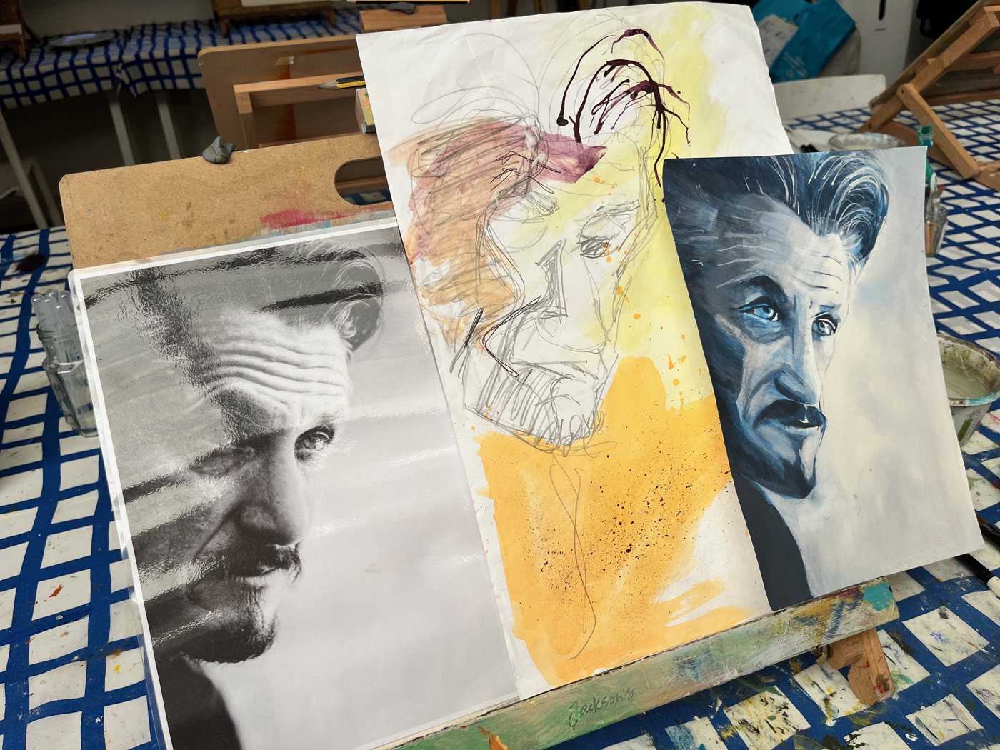 Three portraits on the easel: a black and white study, a loose yellow and pink watercolour face, and a blue painted portrait