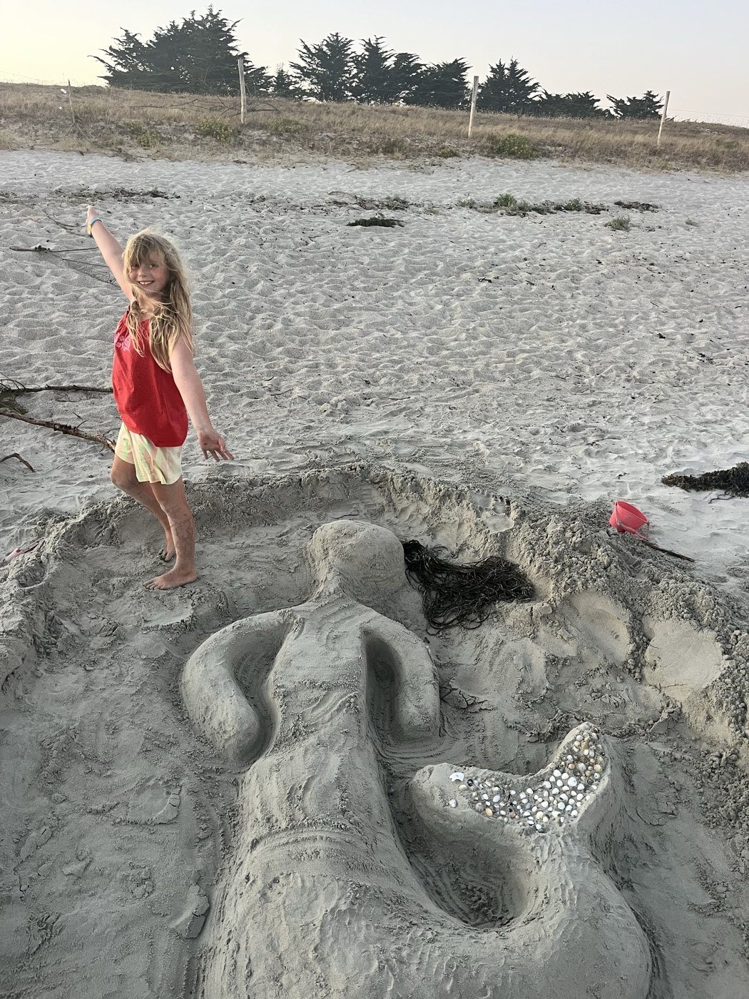 The finished sand mermaid: shells set into the tail, a long sweep of seaweed for hair, Amélie standing beside it