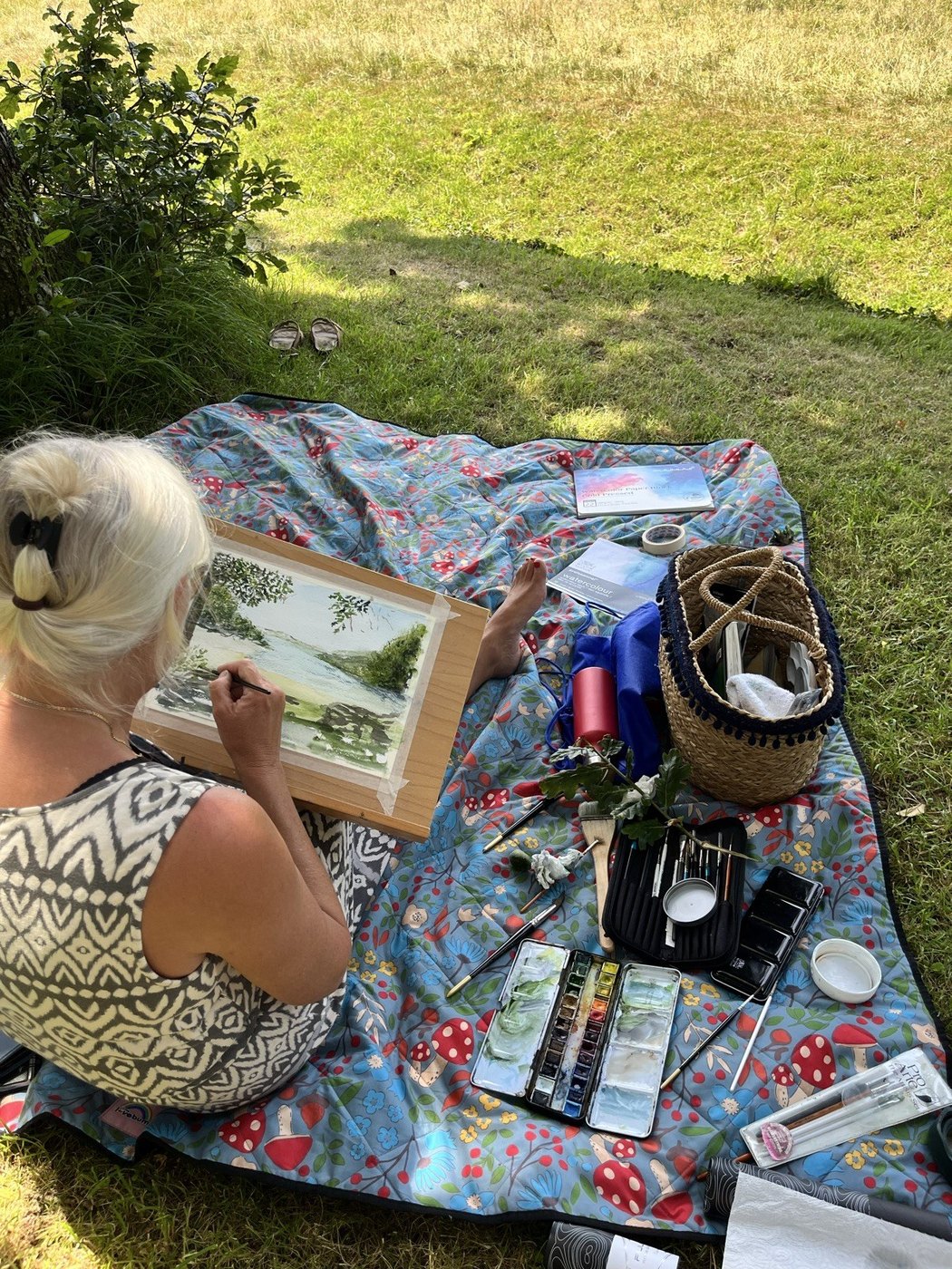 An artist painting on a picnic blanket, kit spread around her